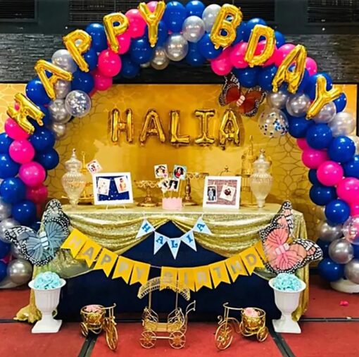 Vibrant birthday party setup featuring a blue, pink, and silver balloon arch with 'Happy Birthday' foil balloon, personalized name balloons, balloon pillars with number foil balloons, a decorated table with 'Happy Birthday' bunting, and 50 balloon bunches around the venue.